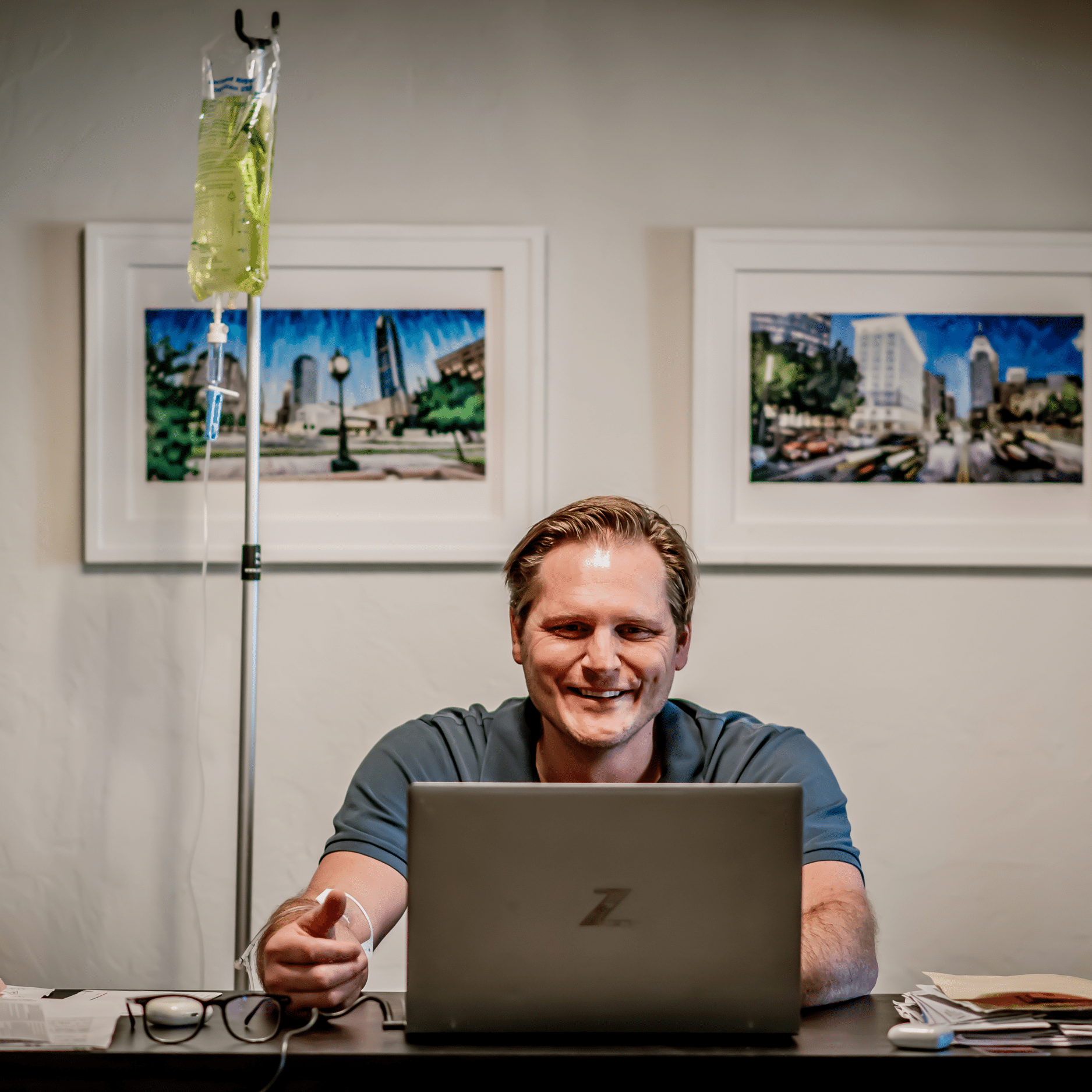 Man in blue shirt working on laptop at office desk with IV therapy stand, city skyline framed artwork on white walls in medical office setting
